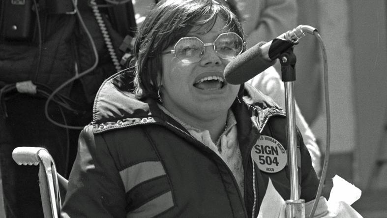 Das historische schwarz-weiße Bild zeigt eine Frau im Rollstuhl, die bei einer Kundgebung ins Mikrofon spricht.