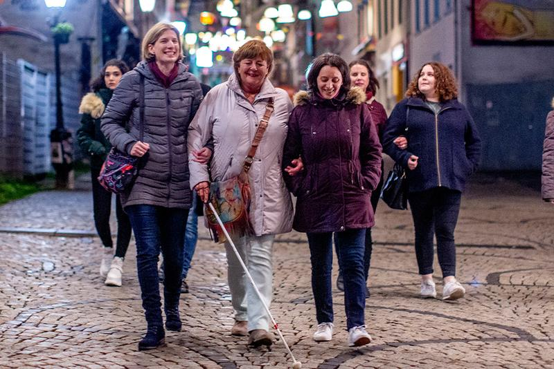 Ein Gruppe Frauen geht durch eine Fussgängerzone. Eine Frau ist sehbehindert und führt ihren Blindenstock.