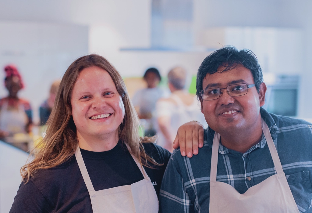 Eine Frau und ein Mann mit Migrationsgeschichte lächeln bei einer Kochveranstaltung in die Kamera.
