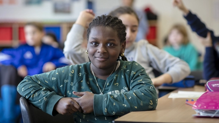 Im Klassenzimmer: Ein Mädchen lächelt in die Kamera, im Hintergrund sitzen SchülerInnen, einige melden sich.