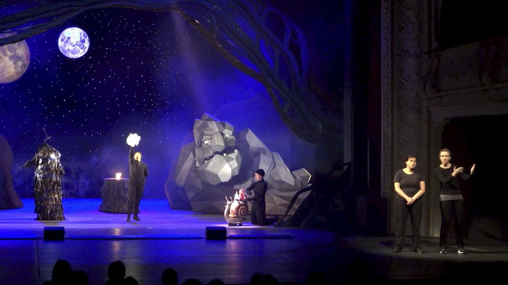 Theaterbühne mit einem Nachthimmel, einem Steinriesen und einem Mann in schwarz, der einen leuchtenden Stern in der Hand hält. Daneben stehen im Scheinwerferlicht zwei Gebärdensprachdolmetscherinnen.