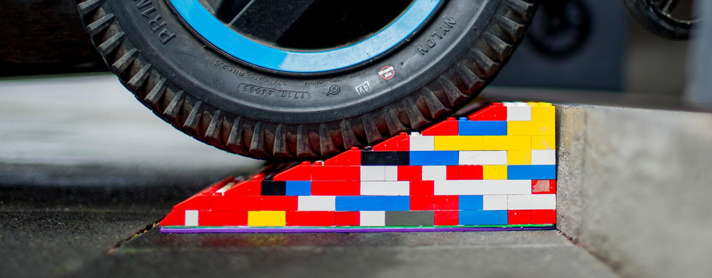 Eine bunte Legorampe liegt vor einer Stufe. Im Anschnitt ist ein Rollstuhl- oder Kinderwagen-Reifen zu sehen, der gerade über die Rampe rollt.