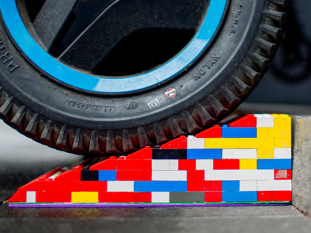 Eine bunte Legorampe liegt vor einer Stufe. Im Anschnitt ist ein Rollstuhl- oder Kinderwagen-Reifen zu sehen, der gerade über die Rampe rollt.