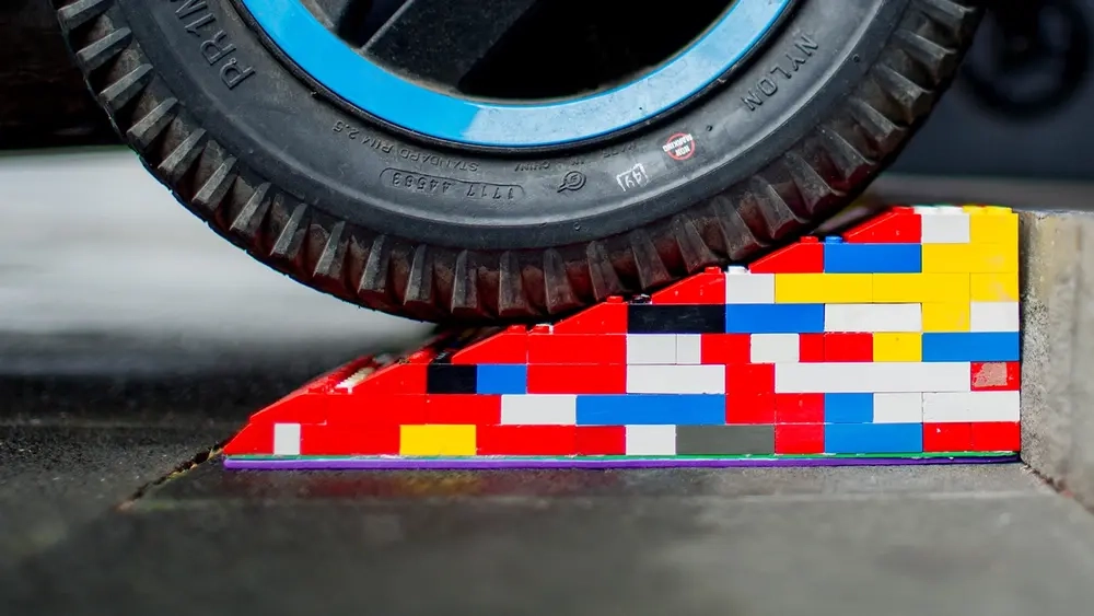 Eine bunte Legorampe liegt vor einer Stufe. Im Anschnitt ist ein Rollstuhl- oder Kinderwagen-Reifen zu sehen, der gerade über die Rampe rollt.