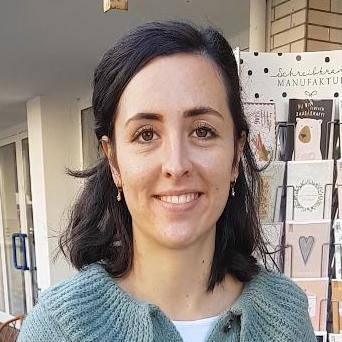 Eine Frau mit dunklem Haar und Strickjacke steht lächelnd vor einer Buchhandlung.