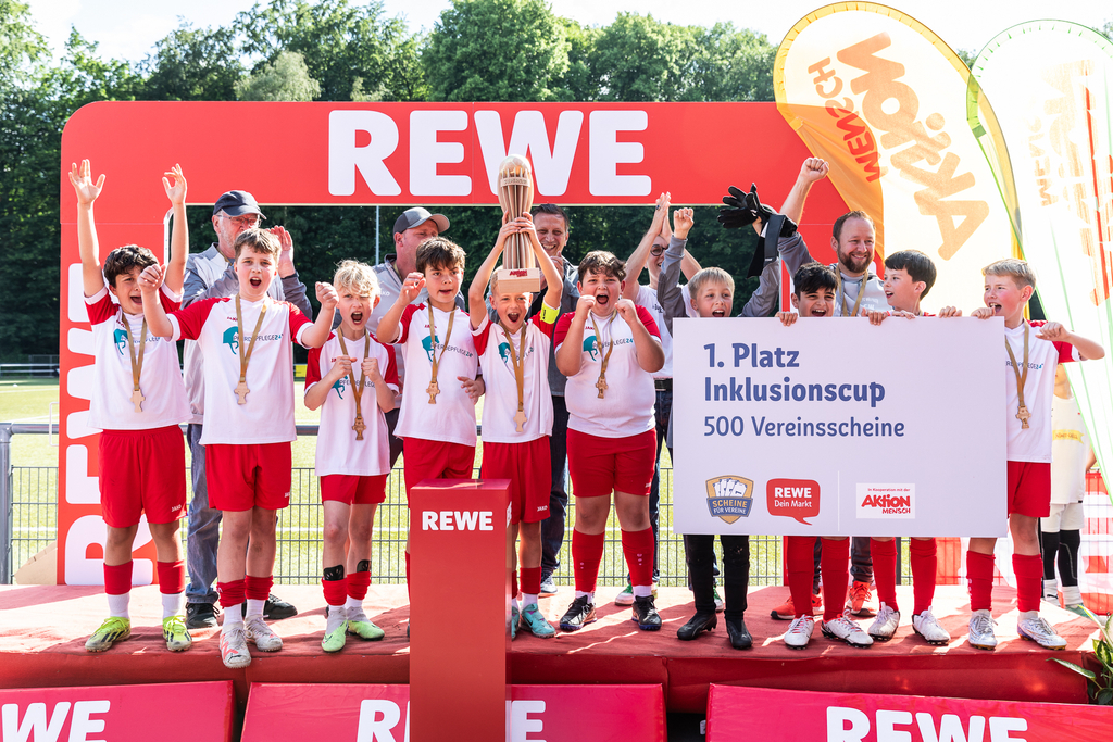 Eine Jungen-Fußballmannschaft feiert auf einem Podest bei der Siegerehrung ihren 1. Platz. Der Kapitän in der Mitte hält den Pokal in die Höhe, drei andere Jungen (etwa 12 Jahre) halten ein Schild in den Händen auf dem steht: "1. Platz Inklusionscup - 500 Vereinsscheine".