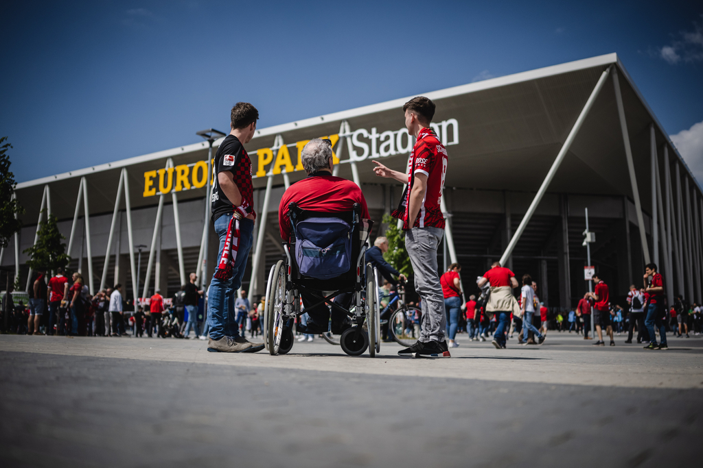 Vor einem Fußballstadion mit dem Schriftzug "Europa Park Stadion" unterhalten sich drei Männer miteinander. Einer der drei sitzt im Rollstuhl. Alle tragen schwarz-rote Fankleidung und schauen zum Eingang des Stadions.