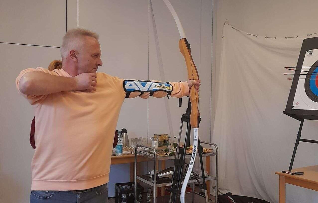 Ein Mann zielt mit einem Bogen auf eine Zielscheibe. Er steht in einem Raum.