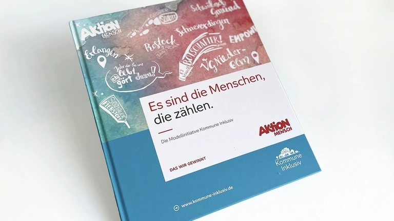 Das Buch "Es sind die Menschen, die zählen" liegt auf einem Tisch. Die obere Hälfte des Einbandes ist mit Zeichnungen verziert , die untere Hälfte ist blau.