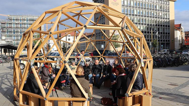 Menschen sitzen unter einer Holzkuppel.