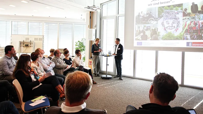 Ein Mann und eine Frau stehen vor einer Gruppe von Menschen und moderieren