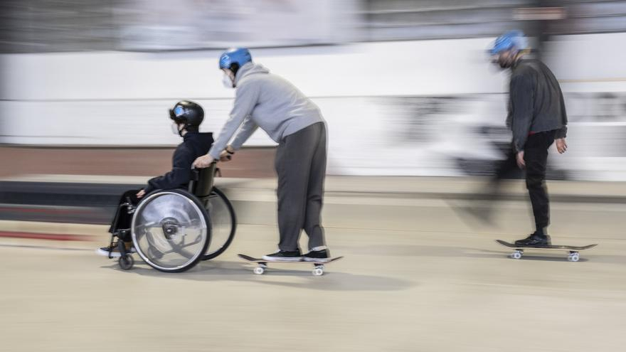 Eine Person mit Helm auf einem Skateboard schiebt eine Person mit Helm im Rollstuhl. Dahinter fährt eine Person mit Helm auf einem Skateboard.