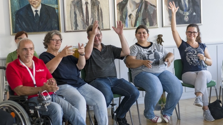 Mehrere Personen mit und ohne Behinderung sitzen in einer Reihe, heben die Hände und lächeln