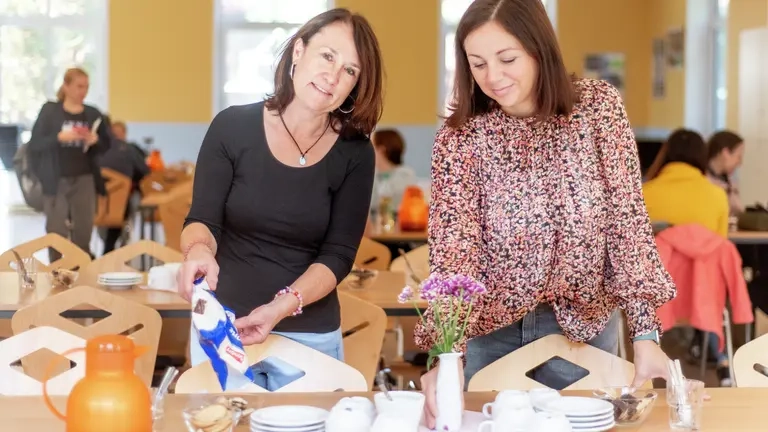 Zwei Frauen bereiten einen Tisch zum Kaffee trinken vor. Im Hintergrund sitzen Menschen an Tischen und unterhalten sich.