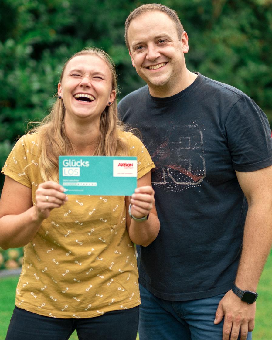 Auf dem Bild lachen zwei Personen fröhlich in die Kamera. Die Frau hält ein "Glücks LOS" von Aktion Mensch in den Händen, während der Mann neben ihr steht und ebenfalls lächelt.