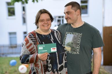 Eine Frau und ein Mann stehen Arm in Arm nebeneinander. Die Frau hält ein Glücks-Los in die Kamera. Der Mann lächelt die Frau verliebt an.