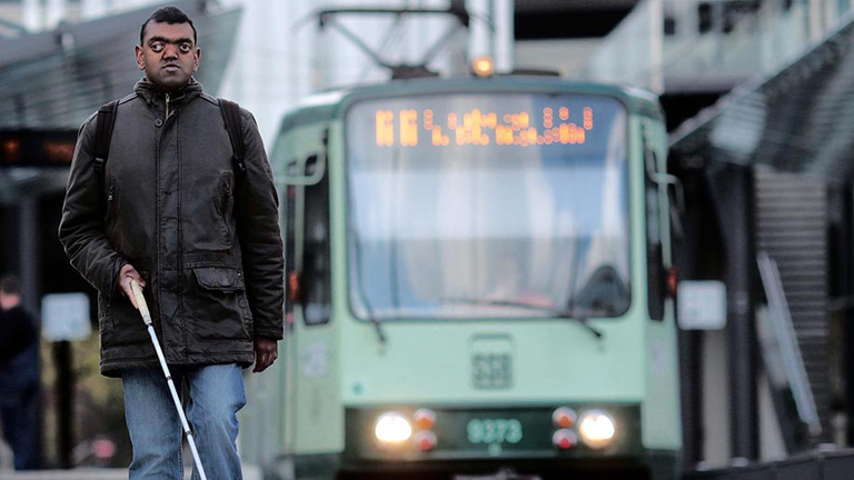 Ein Mann mit Blindenstock geht einen Bahnsteig entlang