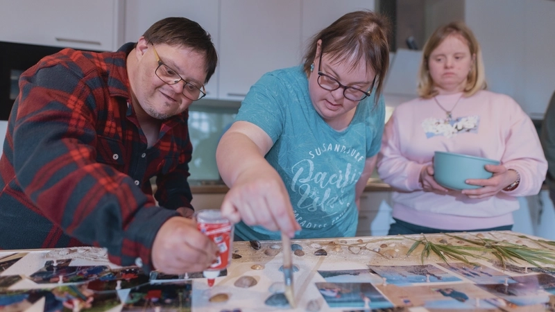Ein junger Mann im Holzfäller-Hemd und zwei junge Frauen stehen in einer Wohnhausküche und basteln an einer Fotocollage. Auf ein großes Brett haben sie Urlaubs-Fotos gepinnt und kleben um sie herum Sand, Muscheln und Schilf. Alle drei haben eine Beeinträchtigung.