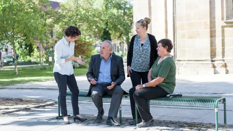 Drei Frauen und ein Mann unterhalten sich im Park auf einer Bank vor einem Gebäude