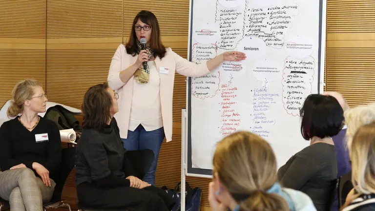 Eine Frau zeigt auf einer Pinnwand und spricht in ein Mikrofon, mehrere Personen hören zu.