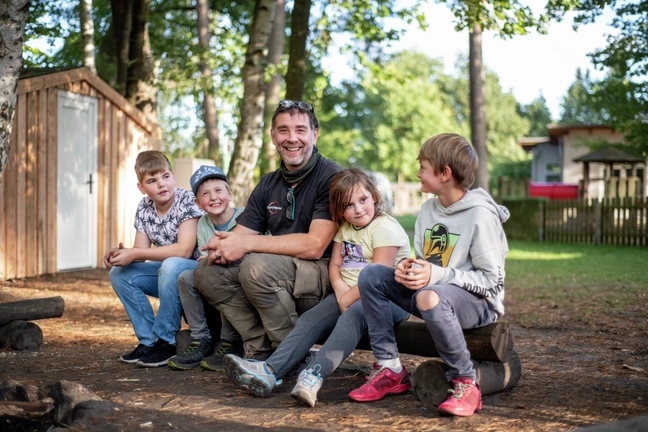 Vier Kinder und ein Erwachsener sitzen auf einem Bank aus einem Baumstamm in einem Park