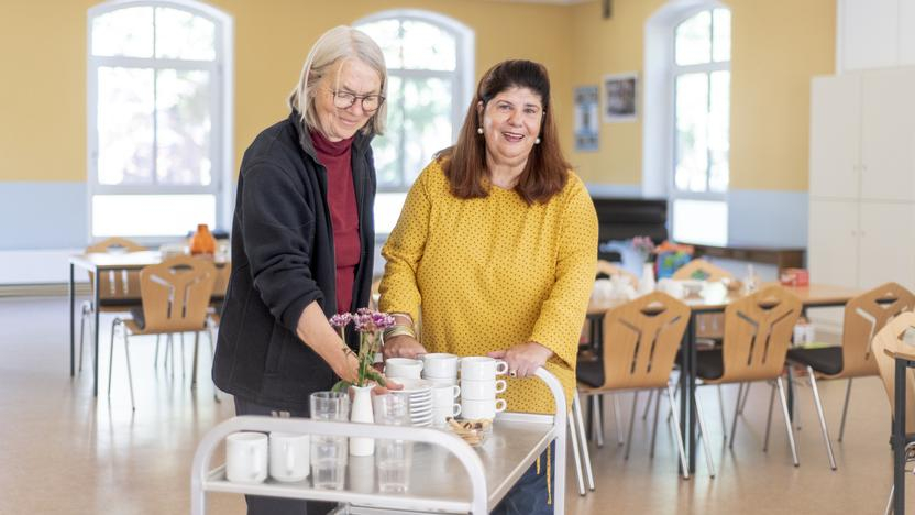 Zwei ältere Frauen bereiten einen Kaffee-Treff vor, sie verteilen Tassen auf Tische.
