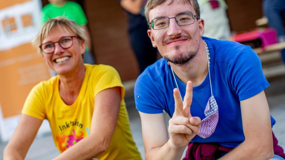 Eine Frau und ein Mann mit geistiger Behinderung sitzen lächelnd nebeneinander. er macht ein Victory-Zeichen
