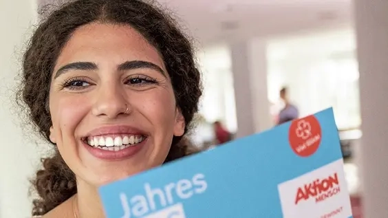 Eine Frau lächelt in die Kamera. In ihrer rechten Hand hält sie ein Jahreslos der Aktion Mensch.