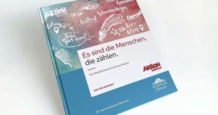 Das Buch "Es sind die Menschen, die zählen" liegt auf einem Tisch. Die obere Hälfte des Einbandes ist mit Zeichnungen verziert , die untere Hälfte ist blau.