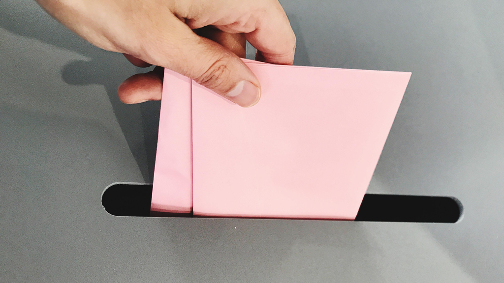 Eine Hand wirft einen rosa Umschlag in eine Wahlurne.