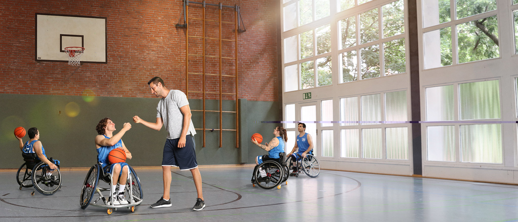 Ein Basketballer im Rollstuhl im blauen Trikot schlägt mit seinem Trainer ein. Im Hintergrund machen sich einige Basketballer*innen im Rollstuhl bereit, auf den Korb zu werfen.