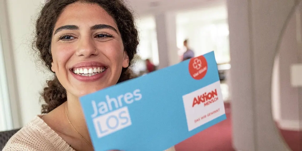 Eine Frau lächelt in die Kamera. In ihrer rechten Hand hält sie ein Jahreslos der Aktion Mensch.