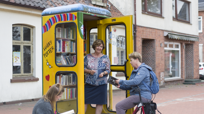 In einer alten Telefonzelle ist jetzt ein Bücherschrank, drei Personen lesen in Büchern.