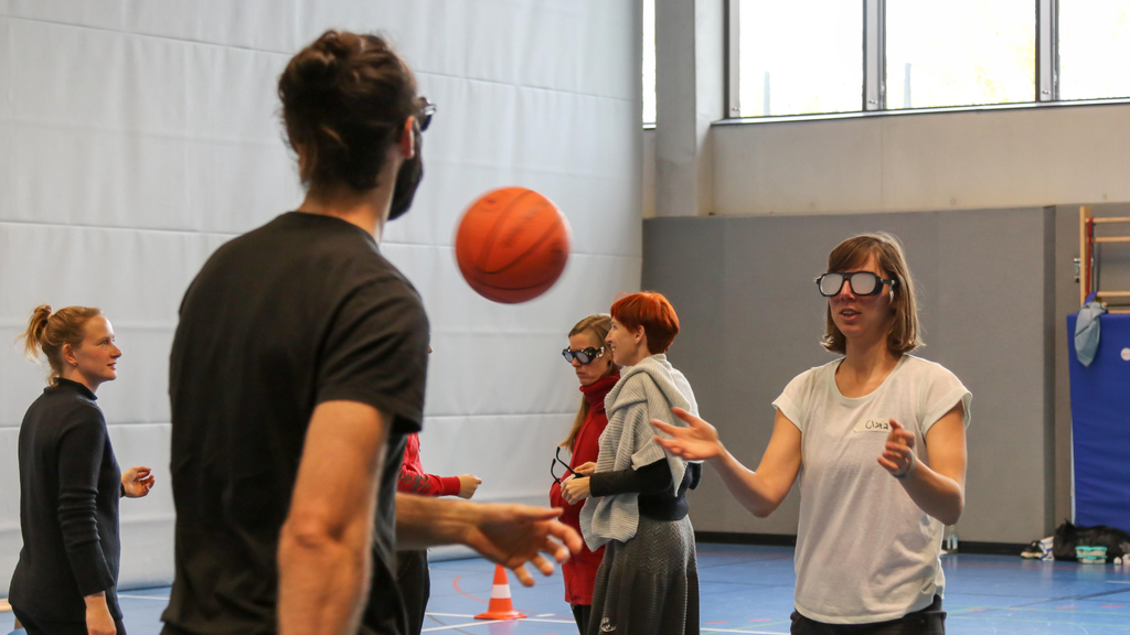 In einer Sporthalle werfen sich Menschen einen Basketball zu. Manche von ihnen tragen eine große Brille mit milchigen Gläsern.