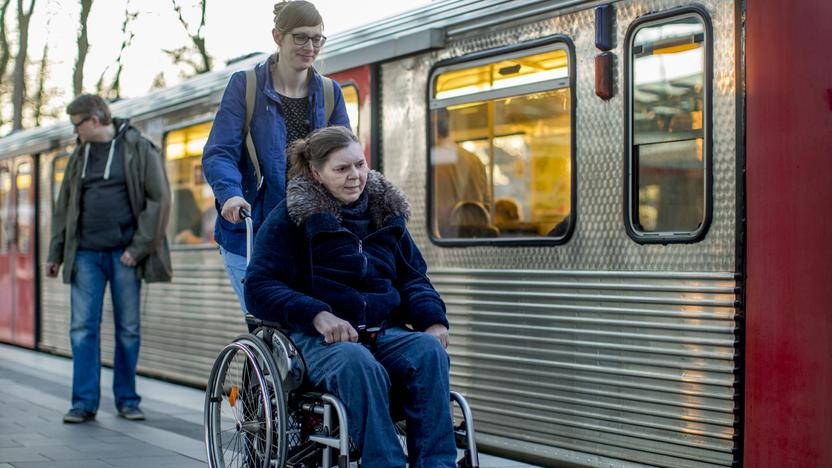 Eine ältere Frau sitzt in einem Rollstuhl, der von einer jüngeren Frau geschoben wird. Die beiden sind an einem Bahnsteig, an dem eine Bahn steht.