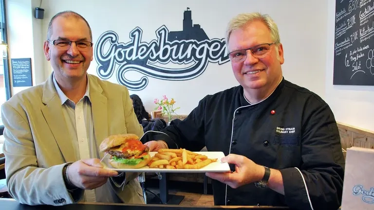Zwei Männer halten einen Teller mit einem Burger in die Kamera.