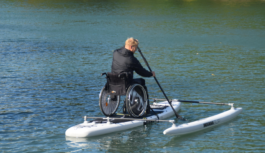 Auf einem besonderen Standup-Paddle, auf dem Wasser, ist ein Mann im Rollstuhl zu sehen, der ein Ruder in der Hand hat.