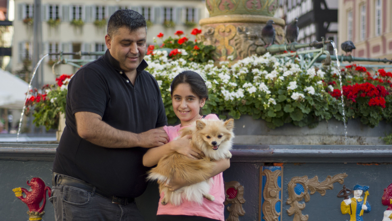 Ein Mädchen hält einen Hund auf dem Arm, der Vater steht daneben, sie stehen draußen vor einem bunt verziertem Brunnen