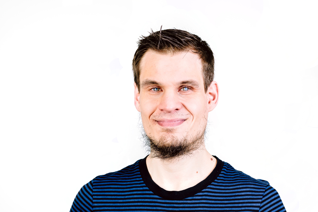 Ein junger Mann mit braunen Haaren und Bart schaut in die Kamera. Er trägt ein schwarz blau gestreiftes T-Shirt.