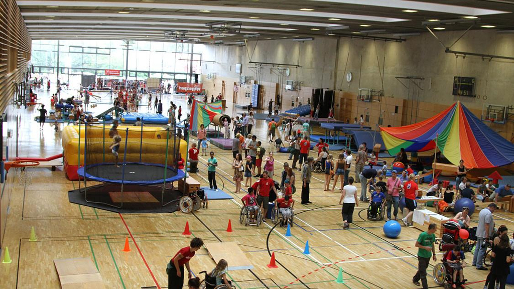 In einer Sporthalle sind viele Stationen aufgebaut, zum Beispiel ein großes Trampolin und ein Zirkuszelt. An den Stationen sind verschiedene Menschen.
