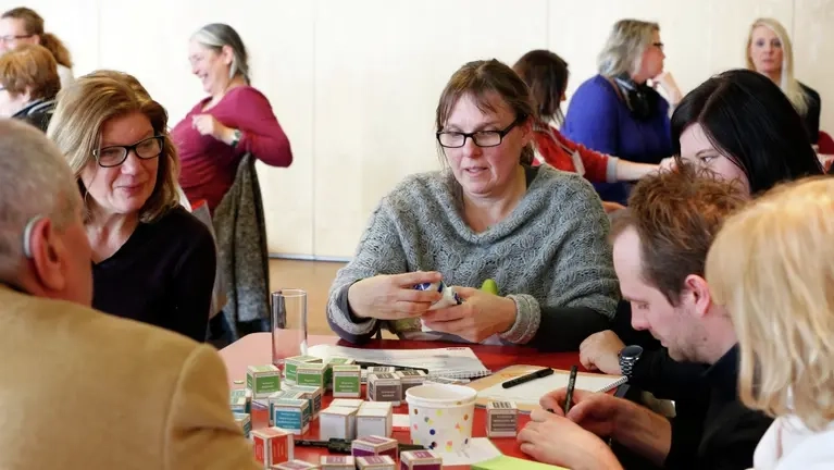 Eine Gruppe von Menschen sitzen an einem Tisch und schauen beschriftete Würfel an.