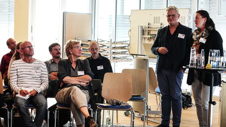 Zwei Menschen stehen an einem Stehtisch und halten einen Vortrag, das Publikum sitzt auf Stühlen hört zu.