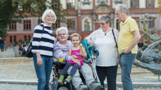 Vier Männer und Frauen, davon eine im Rollstuhl sowie ein Kleinkind vor einer Stadtkulisse.