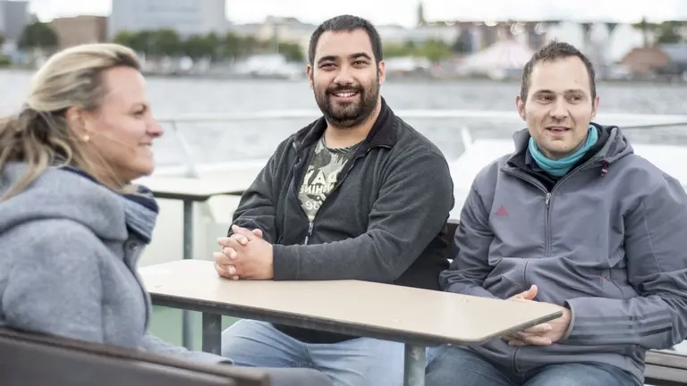 Drei Personen sitzen an einem Tisch, direkt am Meer. Sie sind warm angezogen.