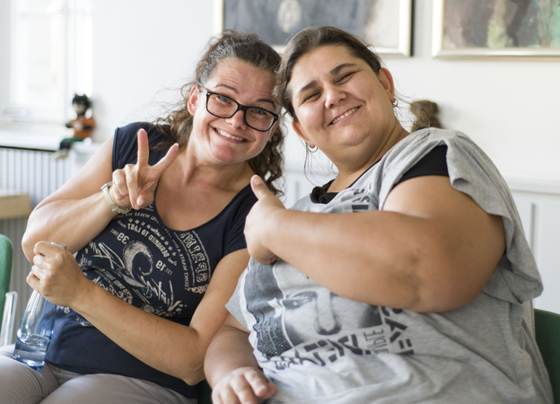 Zwei Frauen lachen in die Kamera, eine zeigt "Daumen hoch", die andere macht ein "Peace"-Zeichen.