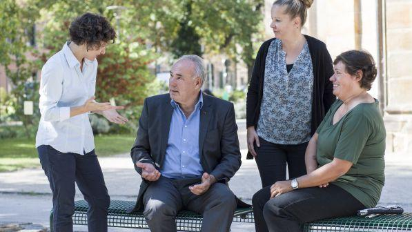 Eine Frau redet mit einem Mann, zwei weitere Frauen hören dem Gespräch zu. Sie sind in einer Grünanlage der Mann und eine Frau sitzen auf einer Bank.