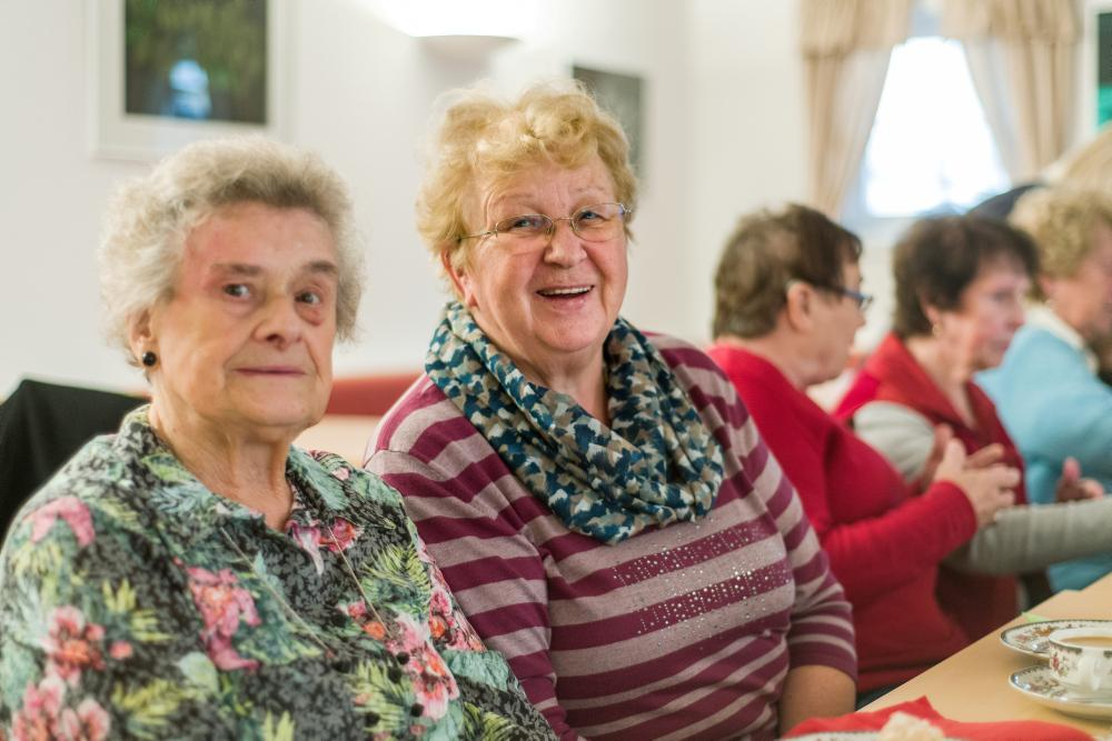 Zwei ältere Damen sitzen am Tisch und lächeln in die Kamera. 