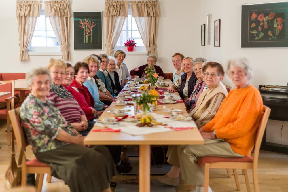 Senior*innen sitzen an einem langen Tisch mit Kaffee und Kuchen. 