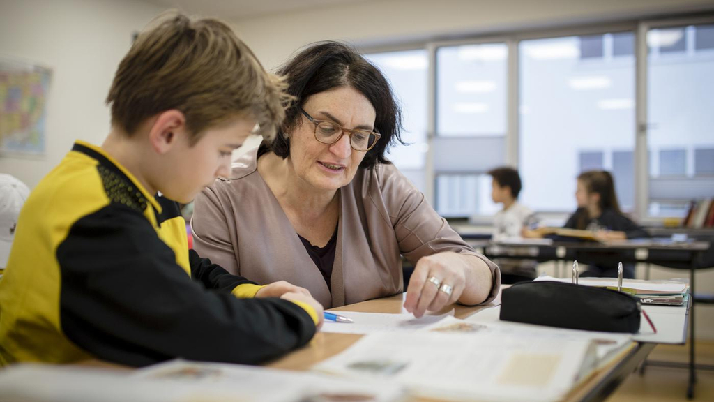 Eine Lehrerin sitzt mit einem Schüler an einem Tisch im Klassenzimmer und erklärt ihm eine Aufgabe.