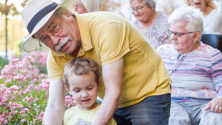 Ein Mann und ein kleines Kind in einem Garten. Im Hintergrund sind eine Seniorin im Rollstuhl und weitere Personen zu sehen.